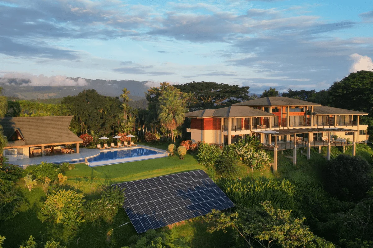 Drone aerial view of a luxury tropical resort with a swimming pool, solar panel array, and surrounding green rainforest hills during golden hour.