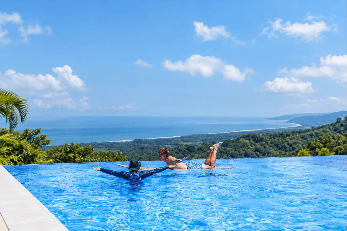 A surf instructor guiding a student on surf stance and pop-up balance technique while lying on a surfboard in a blue infinity pool overlooking the ocean.