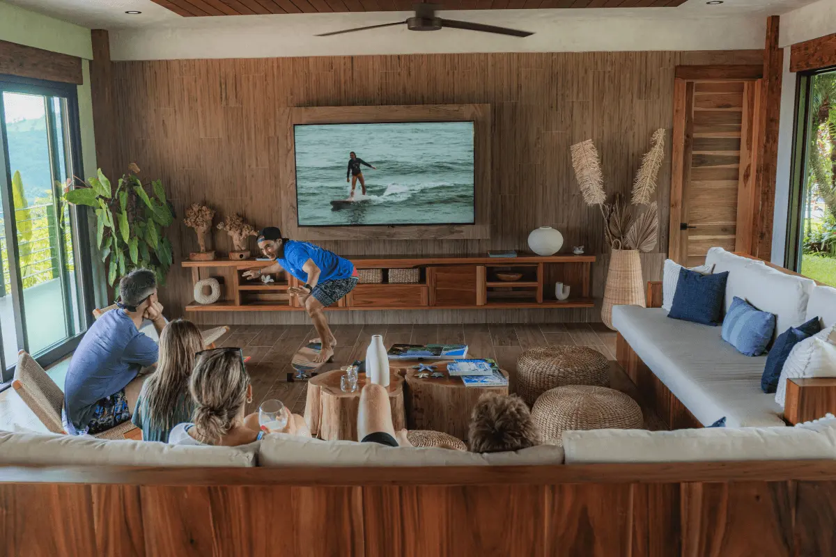 A surf instructor demonstrating surf stance on a skateboard while guests watch and review surfing footage on a large wall TV in a modern lounge.