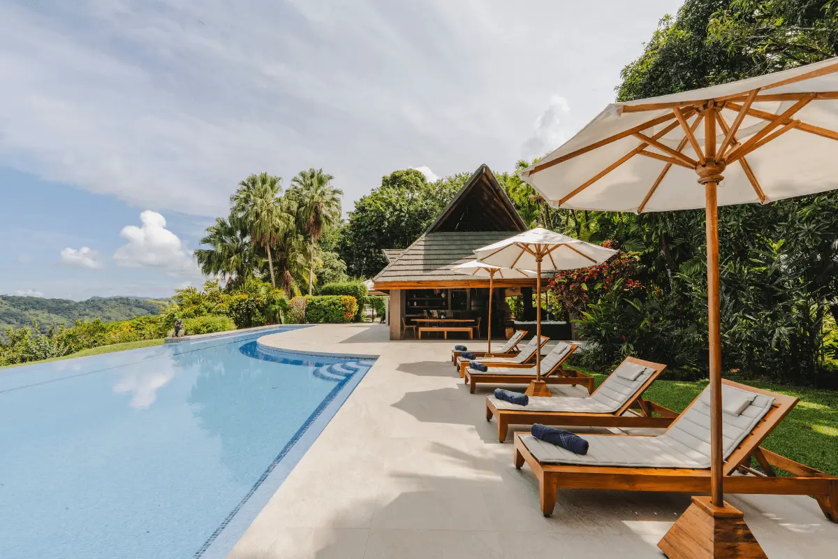 A tranquil resort infinity pool lined with wooden lounge chairs, sun umbrellas, and tropical foliage overlooking lush green hills.