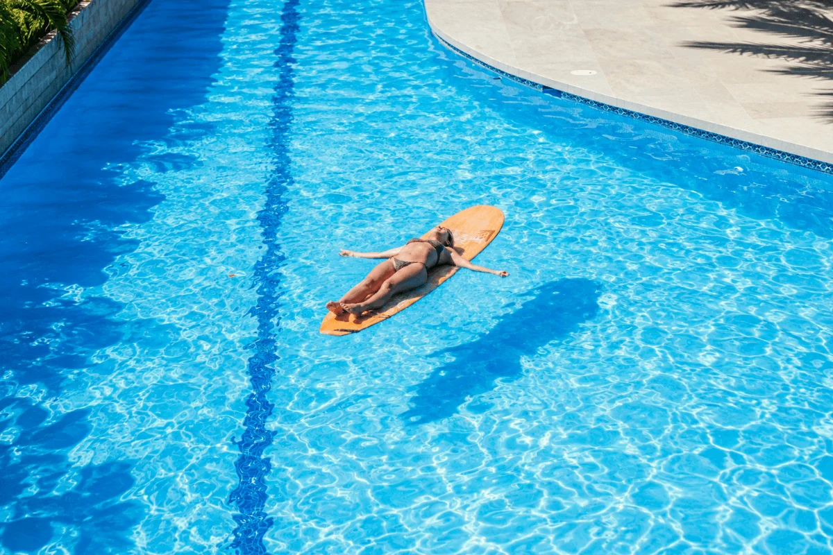 A woman floating peacefully on her back on top of a surfboard in a clear blue swimming pool.