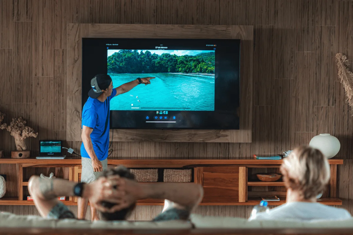 A surf coach pointing at a TV screen showing drone surf footage during a video analysis coaching session with students sitting on a couch.