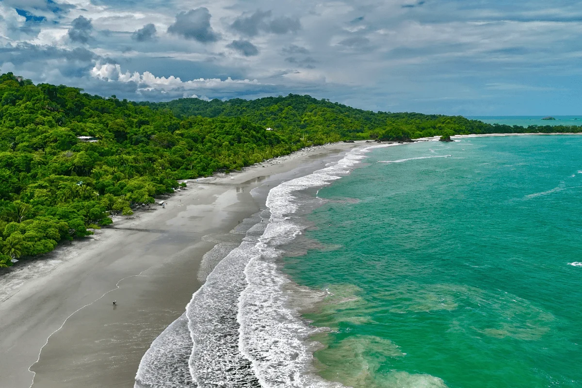 Panoramic drone shot showing rolling ocean waves breaking along a sandy beach bordered by thick tropical rainforest hills in Costa Rica.