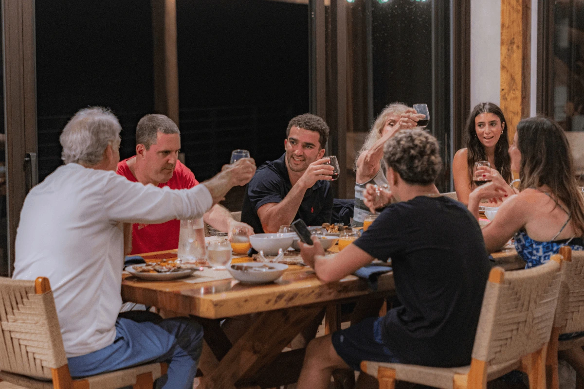 A multi-generational family seated around a large wooden dining table raising their glasses for a toast during dinner at a resort.