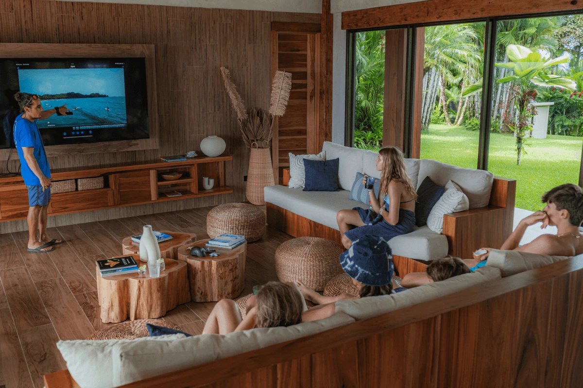 A surf instructor standing by a large television screen pointing out surfing technique to a family gathered on sofas in a resort lounge.