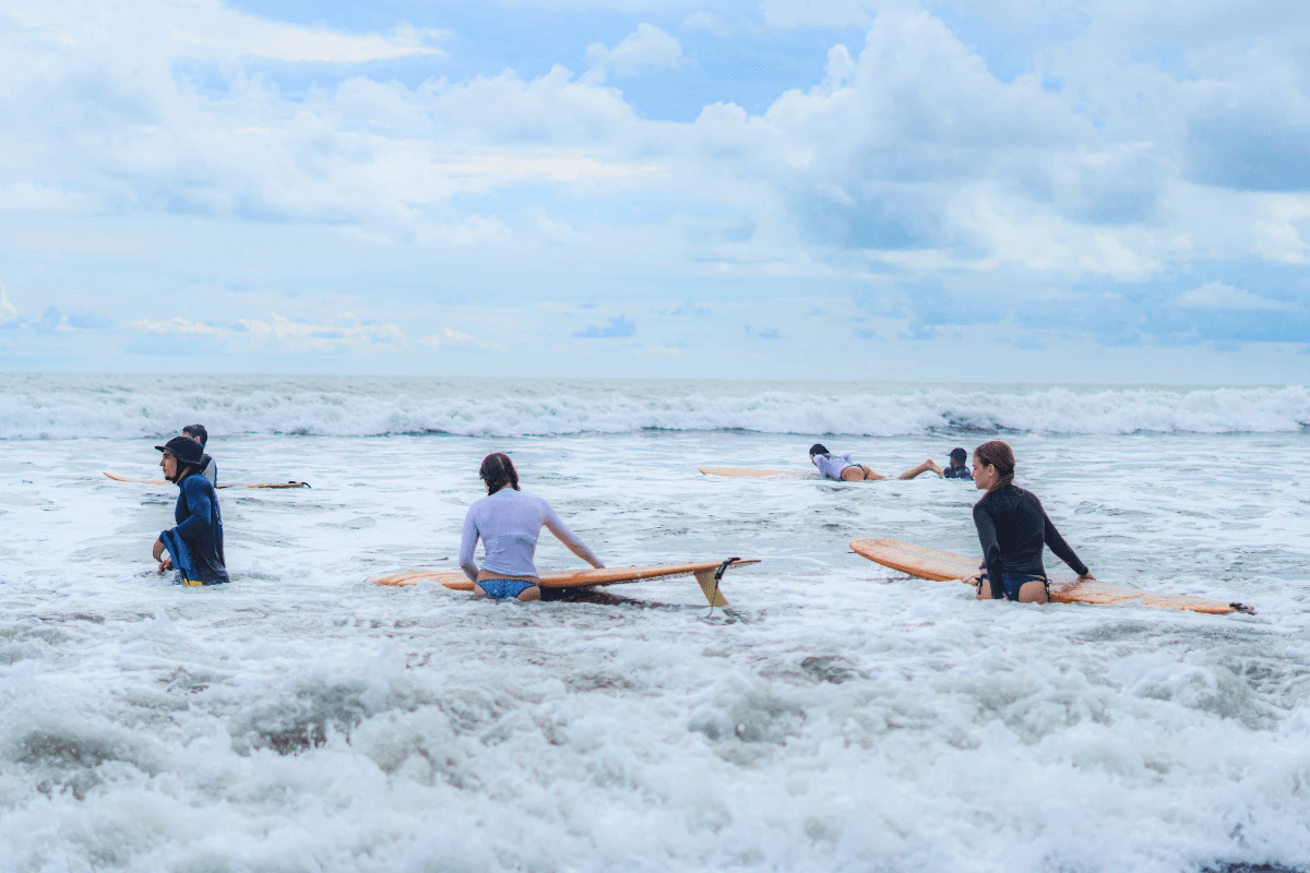 A group of beginners and family members in rashguards walking their surfboards into foaming ocean surf under a cloudy sky.
