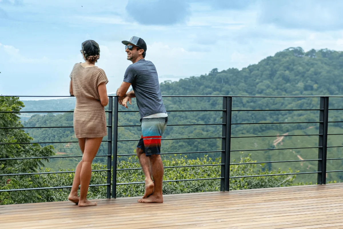 A couple standing on a wooden deck leaning against a railing, enjoying a view of dense green rainforest hills under a cloudy sky.