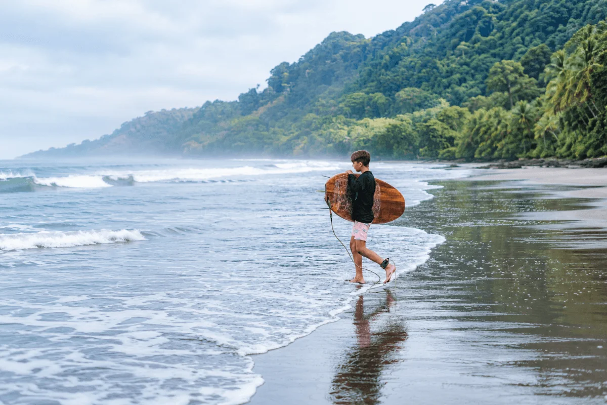 A young boy carrying a wooden surfboard along the tide line on a misty beach with dense jungle mountains in the background
