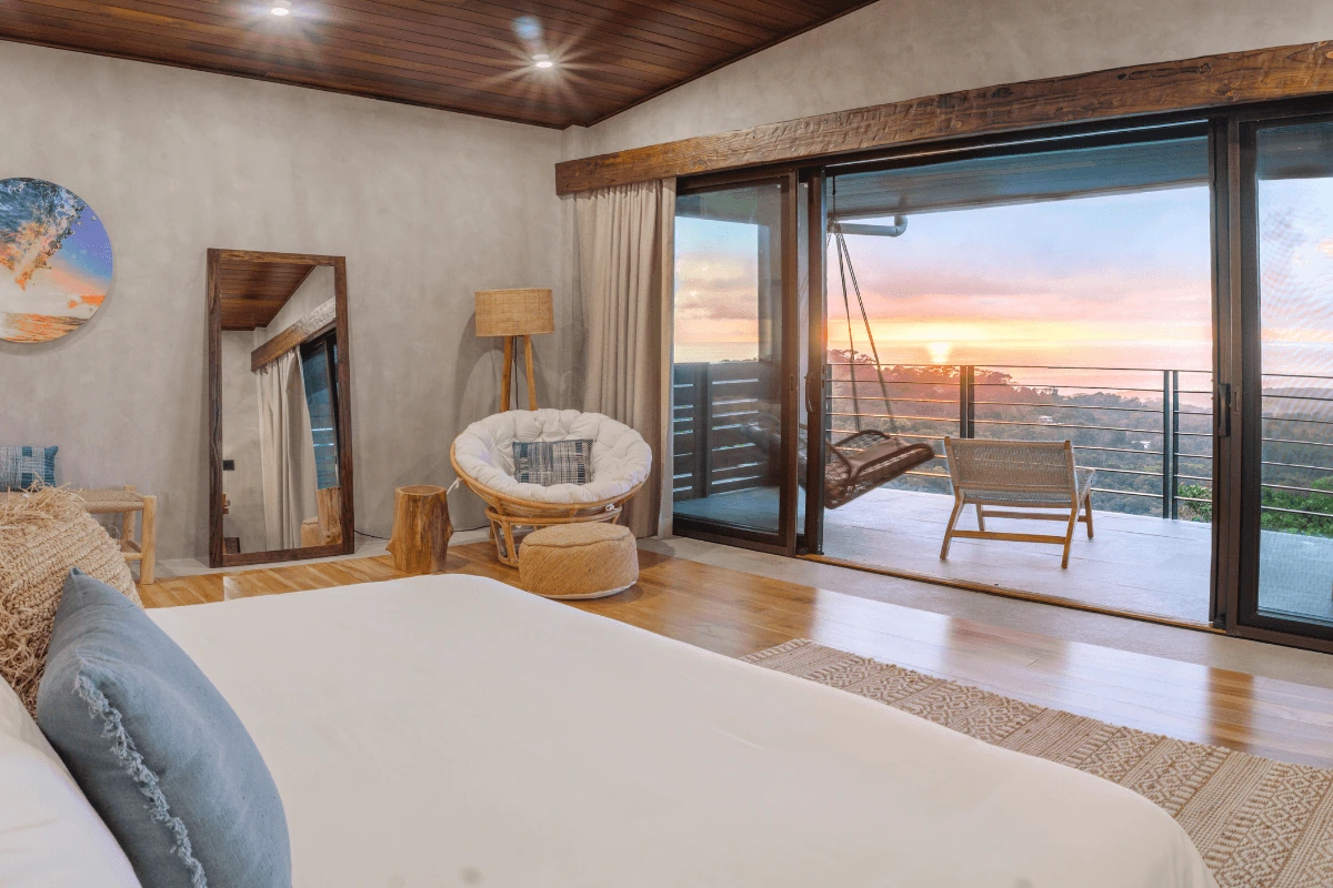 A spacious bedroom with a king bed and glass sliding doors opening to a private balcony with a hanging chair overlooking a mountain sunset.